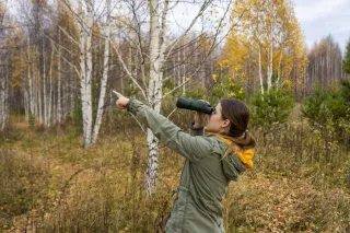 Young woman with binoculars watching birds and pointing in the direction of the bird in the autumn forest