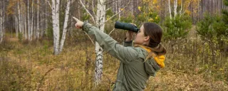 Young woman with binoculars watching birds and pointing in the direction of the bird in the autumn forest