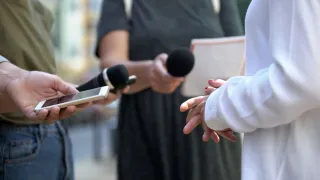 Journalisten befragen unbekannte Person. Mikrofone im Vordergrund. Personen nicht vollständig dargestellt.