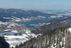 Das Foto zeigt einen Blick von der Bodenschneid auf den Tegernsee. Es ist ein sonniger Wintertag. Die Landschaft wird von Schnee bedeckt.