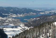 Das Foto zeigt einen Blick von der Bodenschneid auf den Tegernsee. Es ist ein sonniger Wintertag. Die Landschaft wird von Schnee bedeckt.