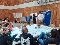 Teilnehmende sitzen um einen Tisch und diskutieren, weitere Teilnehmende stehen vor einer Pinnwand und einem Flipchart und schreiben etwas.