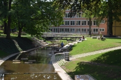 Fluss zwischen Rasenflächen, im Hintergrund eine Brücke und ein Gebäude