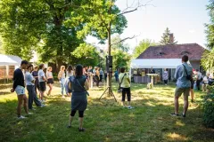 Menschen auf einer Wiese, im Hintergrund ein Pavillon, ein Haus und Bäume