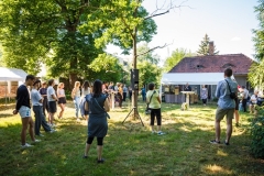 Menschen auf einer Wiese, im Hintergrund ein Pavillon, ein Haus und Bäume