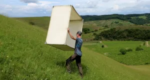 Die Abbildung zeigt die Verwendung des Isolationsquadrats zur Erfassung von Heuschrecken nach der Methode des bundesweiten Insektenmonitorings, ein Mann trägt ein großes Quadrat aus Kunststoff oder Holz über eine grüne Wiese.