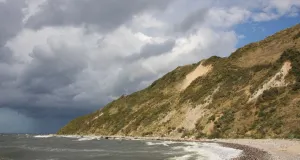 Eine naturbelassene Steilküste mit sichtbaren Hangrutschungen. Das Meer wirkt dunkelgrau. Am Himmel ziehen dunkle Regenwolken auf.