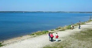 Das Foto zeigt einen See mit Spaziergängern am Ufer.