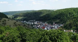 Eine Siedlung in Tallage ist zu sehen. Die angrenzenden Hänge sind weitestgehend bewaldet.