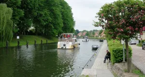 Das Foto zeigt den Fluss Elde in der Stadt Plau am See mit seiner Uferpromenade auf der rechten Flussseite und einen Weiden und Pappelbestand auf der linken Flussseite. Die Elde wir von einer Motoryacht befahren.