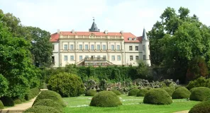 Es sind rundlich geschnittene Buchsgehölze und eine kurz gemähte Rasenfläche zu sehen. In der Mitte des Bildes erscheint ein markantes Schloss.