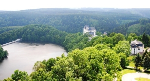 Das Foto zeigt  einen Landschaftsausschnitt des oberen Saaletals mit der Reußischen Residenz Schloss Burgk, einem Teil des Stausees und der Staumauer Burgkhammer. Die umgebende Landschaft ist von geschlossenen Laub- und Nadelwäldern umgeben.