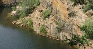 Das Foto zeigt einen aufgelassenen, mit Wasser gefüllten Steinbruch. Eine steile, teils bewachsene, rotbraun schimmernde Felsenwand ragt empor.