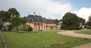 Eine Blumenrabatte säumt eine gemähte Rasenfläche. Im Hintergrund sind ausladende Bäume und das Bergpalais zu sehen.