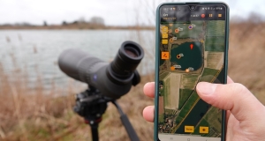 Im Vordergrund sieht man ein Handy mit geöffneter Erfassungs-App, dahinter ein Spektiv am Seeufer