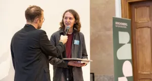 Moderator Lars Ruppel hält ein Mikrofon zu Benedikt Lamberty, der ein Buch in den Händen hält. Im Hintergrund ein Senckenberg-Banner