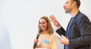 Jessica Holland hält ein Mikrofon und steht neben Lars Ruppel, der mit einer Hand gestikuliert und ein Blatt Papier hält.