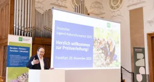 Klement Tockner an weißem Rednerpult in einem prunkvollen Saal mit großer Orgel im Hintergrund. Hinter ihm Leinwand mit Projektion zum Deutschen Jugend-Naturschutzpreis 2025 und ein BfN Banner.