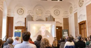 Das Publikum sitzt in einem prunkvollen Saal mit Stuckdecke und schauen auf eine Leinwand, auf der Cornelia Funke per Liveschalte zu sehen ist.