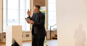 Lars Ruppel trägt schwarzen Anzug und hält ein Tablet, von dem er mit erhobenem Zeigefinger abliest