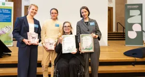 Vier junge Frauen lächeln in die Kamera, eine von ihnen sitzt im Rollstuhl. Sie halten Urkunden und Bücher in den Händen.