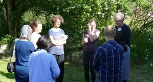 Das Bild zeigt eine kleine Gruppe Teilnehmende, teilweise mit Kaffeetassen in der Hand, die kreisförmig zusammen an einem sonnigen Platz im Grünen stehen und sich unterhalten. 