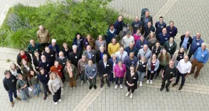 Das Bild zeigt eine Gruppe von etwa 60 Personen, die nah nebeneinander auf einer gepflasterten Fläche neben einem Pflanzbeet stehen und nach oben in die Kamera blicken. Das Foto wurde aus der Vogelperspektive aufgenommen. 
