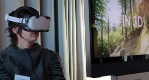 Eine junge Frau mit dunklem Pullover hat ein weißes Virtual Reality Headset auf und sitzt neben einem großen Fernsehbildschirm. Dieser ist in Teilen an der rechten Bildseite zu sehen.