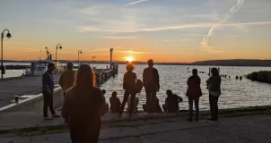 Im Bild sind im Gegenlicht schemenhaft Personen zu sehen, manche sitzend, manche stehend, die in geselliger Runde am Hafenbecken den weit fortgeschrittenen Sonnenuntergang beobachten.
