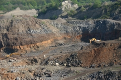 Auf dem Foto ist ein Steinbruch abgebildet. Er zerschneidet die umliegende grüne Landschaft, zwei Bagger bewegen Gestein.