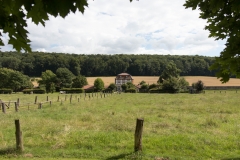 Zwischen eingezäunten Wiesen führt ein geradliniger Weg zu einem Fachwerkhaus. Am Horizont erstreckt sich ein bewaldeter Hang.