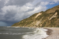 Eine naturbelassene Steilküste mit sichtbaren Hangrutschungen. Das Meer wirkt dunkelgrau. Am Himmel ziehen dunkle Regenwolken auf.