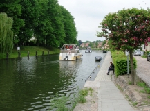 Das Foto zeigt den Fluss Elde in der Stadt Plau am See mit seiner Uferpromenade auf der rechten Flussseite und einen Weiden und Pappelbestand auf der linken Flussseite. Die Elde wir von einer Motoryacht befahren.