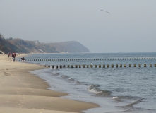 Das Foto zeigt eine Küstenlinie auf Usedom an einem trüben Tag. Am Strand sind mehrere Spaziergänger unterwegs. Im Vordergrund sind Wellenbrecher aus Holzpfosten zu sehen.