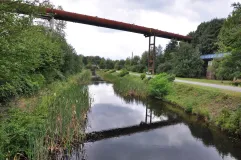 Zu sehen ist ein geradliniger, renaturierter Abschnitt der alten Emscher. Entlang der Ufer wachsen partiell Röhrichte. Eine alte Rohrleitung überquert das Gewässer.