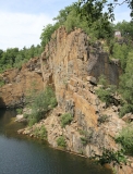 Das Foto zeigt einen aufgelassenen, mit Wasser gefüllten Steinbruch. Eine steile, teils bewachsene, rotbraun schimmernde Felsenwand ragt empor.