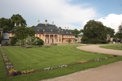 Eine Blumenrabatte säumt eine gemähte Rasenfläche. Im Hintergrund sind ausladende Bäume und das Bergpalais zu sehen.