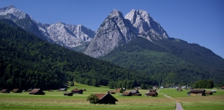 Das Foto zeigt das Wettersteingebirge mit der Alpspitze und dem Waxensteinkamm. Im Tal sind einzelne Wirtschaftsgebäude auf einer Grünlandfläche verteilt.