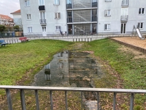 Vor einem mehrgeschossigen Haus befindet eine gewölbte Rasenfläche, die teilweise mit Regenwasser gefüllt ist. An die Rasenfläche grenzt rechts ein Spielplatz mit einer Rutsche an