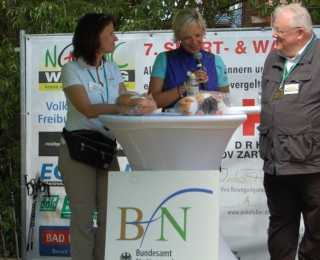 Die Parlamentarische Staatssekretärin Rita Schwarzelühr Sutter (links), in der Mitte eine Moderatorin und rechts Dr. Alfred Herberg als Vertreter der BfN-Hausleitung an einem Stehtisch am Informationsstand des BfN vor einer Werbetafel mit Sponsoren und Partnern der Veranstaltung.