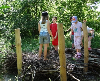 Das Foto zeigt vier spielende Kinder auf einen als Spielelement aufgeschichteten Reisighaufen in einem waldbestockten Naturerfahrungsraum.