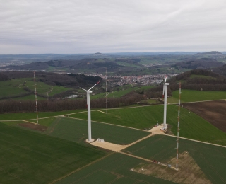 Drohnenaufnahme mit Blick auf zwei Windenergieanlagen samt Messmasten in der Agrarlandschaft