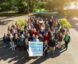 Teilnehmende des MAB Jugendforums halten ein Plakat mit dem Logo des MAB Jugendforums in den Händen und stehen im Kreis um einen Brunnen.