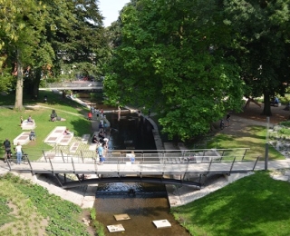 Brücke über einem Fluss, daneben Wiesen mit Bäumen, Sitzelementen und Menschen