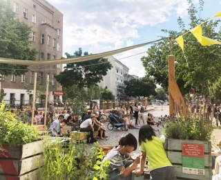Eine Wimpelkette, bepflanzte Hochbeete und spielende Kinder im Vordergrund, viele Menschen sitzen auf einer gesperrten Straße zwischen Häusern im Hintergrund