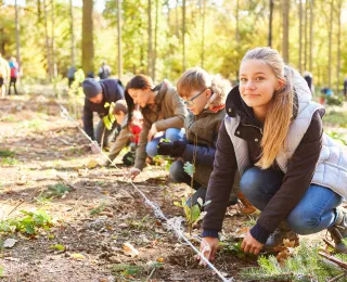 Eine Gruppe Jugenlicher kniet auf dem Waldboden und pflanzt Bäume. Ein Mädchen im Vordergrund des Bildes blickt lächelnd in die Kamera.