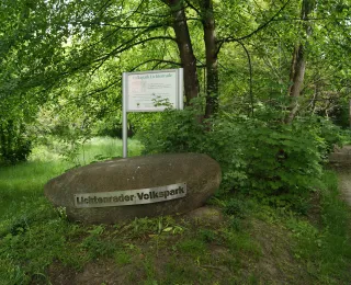 Großer Stein zwischen Bäumen und anderen Pflanzen, ausgestattet mit Infoschild und Beschriftung