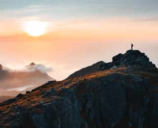 Die Abbildung zeigt einen Bergsteiger bei Sonnenuntergang auf dem Gipfel eines Berges in Norwegen (Lofoten)