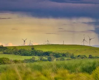 Grüne Hügellandschaft mit mehreren Windenergieanlagen und Strommasten im Hintergrund. Über dem Horizont ziehen Regenwolken auf. Im Vordergrund Wiesen und Bäume.