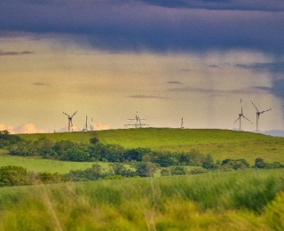 Grüne Hügellandschaft mit mehreren Windenergieanlagen und Strommasten im Hintergrund. Über dem Horizont ziehen Regenwolken auf. Im Vordergrund Wiesen und Bäume.
